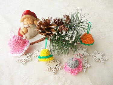 Nikolausfigur neben einem schneebedeckten Tannenzweig; davor vier bunte gehäkelte Anhänger und weiße Schneeflocken-Dekore.
