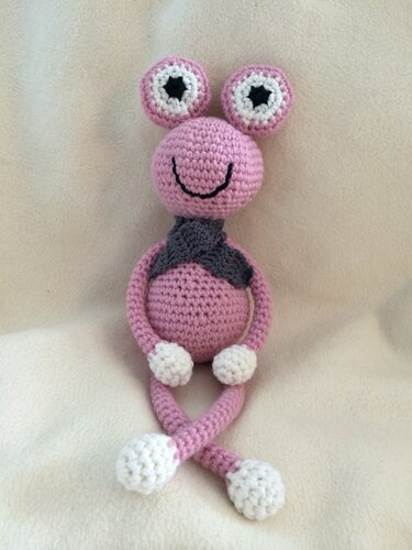 Rosa gehäkelte Amigurumi-Figur mit zwei Augenstielen, lächelndem Gesicht, grauem Schal und gekreuzten Beinen