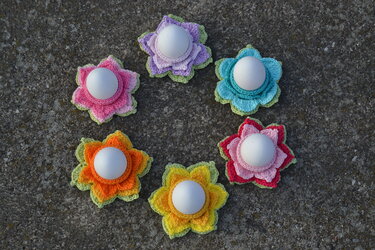 Sechs bunte, gehäkelte Blumen‑Eierwärmer mit weißen Eiern auf grauem Untergrund.