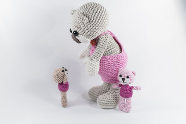 Drei gehäkelte Amigurumi-Figuren: großer hellgrauer Bär mit rosa Latzhose und zwei kleinere Figuren auf weißem Hintergrund