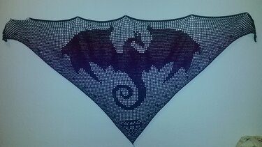 Dreieckiges gehäkeltes Tuch in dunkler Farbe mit Drachenmotiv im Filetgitter an einer Wand