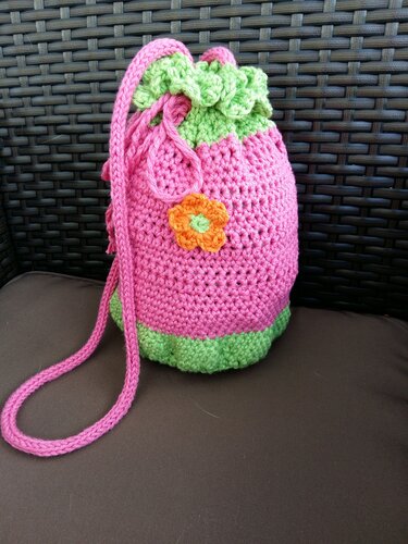 Gehäkelte pink-grüne Beuteltasche mit orangefarbener Blumenapplikation vor dunklem Geflecht