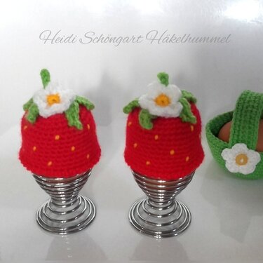 Zwei rote, gehäkelte Erdbeer-Eierwärmer mit grünen Blättern und weißen Blüten auf spiralförmigen Metall-Eierhaltern; rechts ein kleiner grüner Häkelkorb mit Eiern.