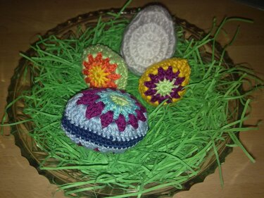 Mehrere eiförmige Häkelmotive in verschiedenen Farben auf grünem Dekogras in einer Glasschale