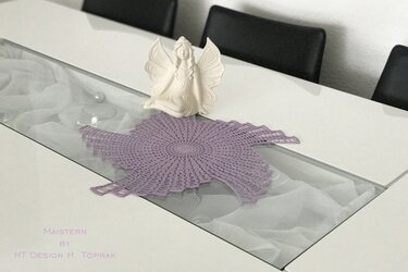 Lavendelfarbenes, sternförmig gehäkeltes Deckchen auf einem Glastisch neben einer Engelsfigur.