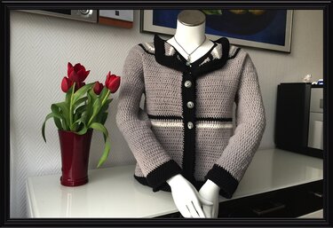 Hellgraue gehäkelte Damenjacke mit vier Knöpfen, schwarzen Bündchen und hellem Taillenstreifen auf einer Schaufensterpuppe.