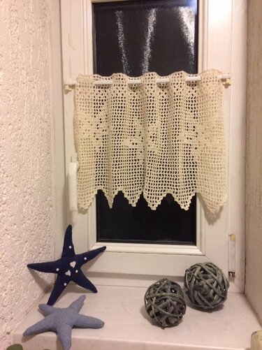 Handgehäkelter, cremefarbener Vorhang mit Muschelmuster an kleinem Fenster; Stoff-Seesterne und geflochtene Kugeln auf der Fensterbank.
