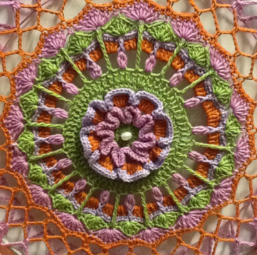 Gehäkeltes rundes Mandala mit Blumenmotiv, Spitzenmuster und weißer Perle in der Mitte.