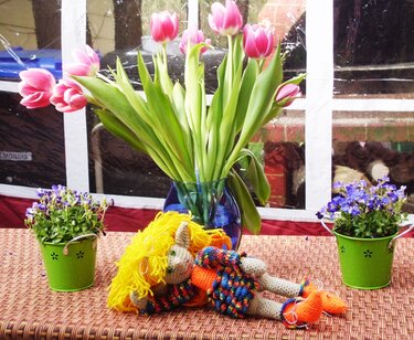 Gehäkelte Amigurumi-Puppe mit gelbem Haar und buntem Outfit, liegend zwischen Tulpen und Topfpflanzen