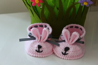 Zwei rosa gehäkelte Babyschuhe mit Hasenohren vor grünem Filzkorb mit Blumen