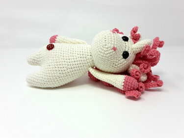 Gehäkeltes weißes Einhorn-Amigurumi mit rosa Lockenmähne, schwarzen Augen und Knopf, seitlich liegend