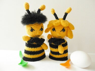 Zwei gehäkelte Bienen-Eierwärmer in Gelb und Schwarz mit grimmigen Gesichtern, neben einem Ei und kleinen Löffeln.