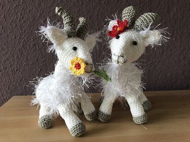 Zwei gehäkelte weiße Ziegen-Amigurumi mit grauen Hörnern und gehäkelten Blumen auf Holz