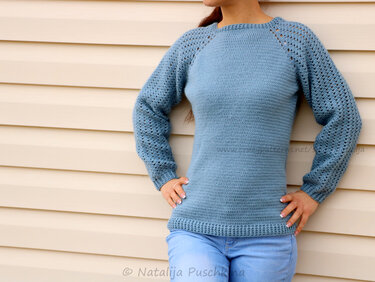 Hellblauer gehäkelter Damenpullover mit langen Raglanärmeln im Lochmuster, stehend vor einer beigefarbenen Wand.