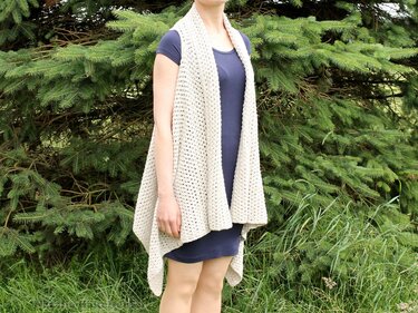 Person trägt helle, lange gehäkelte Weste über dunklem Kleid vor Tannenzweigen