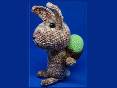 Gehäkelter braun-beiger Hase mit schwarzem Auge, trägt ein grünes Ei in einem braunen Korb, vor blauem Hintergrund.
