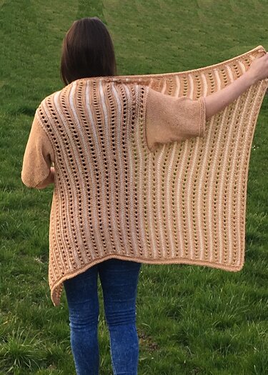 Rückansicht einer Person, die einen beigefarbenen, gestrickten Überwurf mit vertikalem Lochmuster und hellen Streifen trägt, stehend auf einer Wiese.