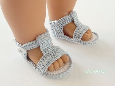 Nahaufnahme hellblauer gehäkelter Babysandalen mit Riemen und weißem Knopf an den Füßen einer Babypuppe.