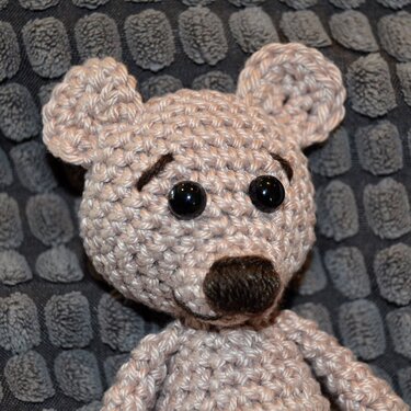 Beigefarbener gehäkelter Teddybär mit schwarzen Augen und brauner Nase vor strukturiertem grauen Hintergrund.