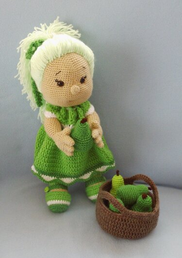 Eine gehäkelte Amigurumi-Puppe in grünem Kleid und mit hellem Zopf, die eine Birne hält. Daneben steht ein Korb mit weiteren gehäkelten Birnen.