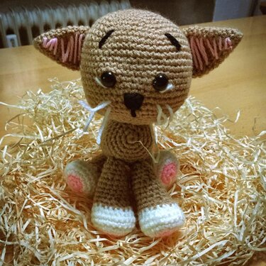 Braune gehäkelte Amigurumi-Katze mit weißen Pfoten und rosa Innenohren, sitzt auf Holzwolle