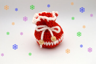 Handgehäkeltes rotes Säckchen mit weißen Verzierungen, weißem Zugband mit zwei Holzperlen, vor hellem Hintergrund mit bunten Schneeflocken.