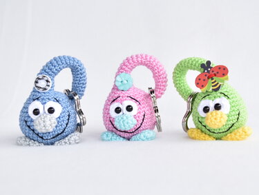 Three crocheted keychains on a white background: a blue one with a checkered button, a pink one with a flower button, and a green one with a ladybug embellishment.