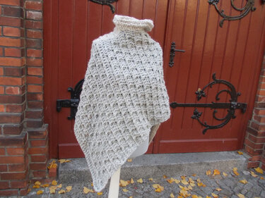 Cremefarbener gehäkelter Poncho mit hohem Rollkragen auf Schaufensterpuppe vor Holztür