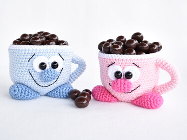Two crocheted cup-shaped holders with smiling faces, one blue and one pink, filled with round chocolate-covered candies
