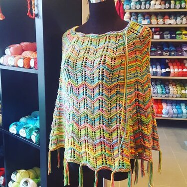 Mehrfarbiger gestrickter Poncho mit Zickzack-Ajourmuster auf schwarzer Schaufensterpuppe vor Garnregal