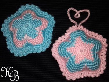 Zwei gehäkelte Sterne in Rosa und Hellblau auf dunklem Hintergrund, einer mit herzförmiger Schlaufe