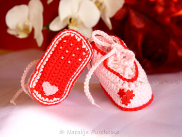 Nahaufnahme zweier gehäkelter Babyschuhe in Rosa und Rot mit Herz- und Blumenapplikationen.