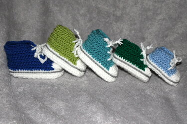 Reihe aus fünf gehäkelten Babyschuhen in Dunkelblau, Hellgrün, Türkis, Dunkelgrün und Hellblau mit weißen Sohlen auf grauem Stoff
