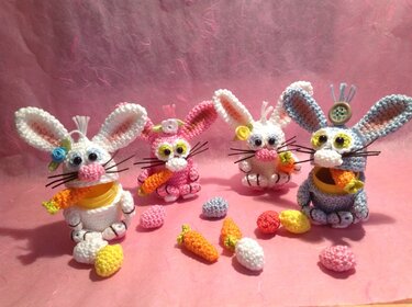 Vier handgehäkelte Amigurumi-Osterhasen in Weiß, Rosa und Hellblau mit kleinen gehäkelten Möhren und bunten Eiern vor rosafarbenem Hintergrund