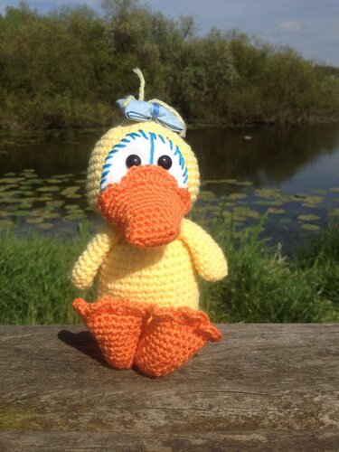 Gehäkeltes gelbes Küken mit orangefarbenem Schnabel sitzend auf einem Holzstamm vor einem Teich mit Seerosen.