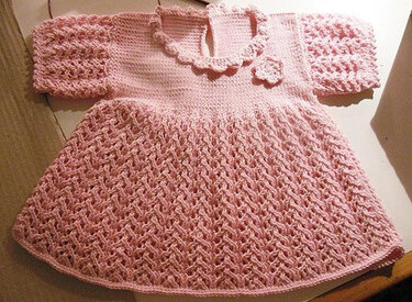 Rosafarbenes, gestricktes Babykleid mit kurzen Ärmeln, gewelltem Kragen, Blumenapplikation und Strukturmuster am Rock.