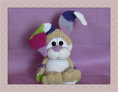 Sitzender Amigurumi-Hase in Beige und Weiß mit bunten Patchwork-Ohren vor violettem Hintergrund