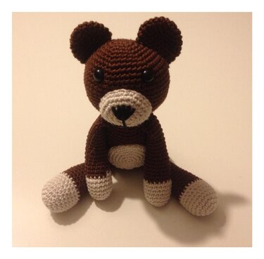Sitzender brauner Amigurumi-Teddybär mit heller Schnauze, Bauch und Pfoten vor hellem Hintergrund.