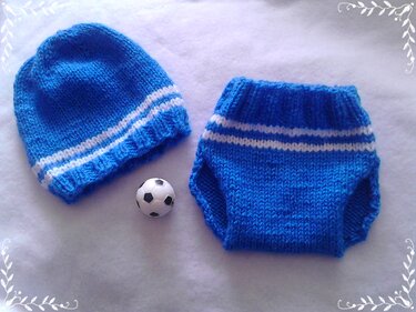 Gestricktes blaues Baby-Set mit weißen Streifen: Mütze und Windelhose, daneben ein kleiner Fußball auf hellem Untergrund.