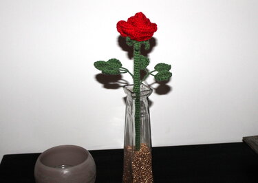 Gehäkelte rote Rose mit grünen Blättern in schlanker Glasvase mit goldfarbenem Dekogranulat vor weißem Hintergrund.