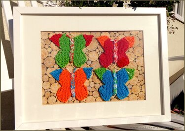 Weißer Bilderrahmen mit vier gestrickten Schmetterlingen in Grün, Rot, Orange und Blau auf Holzscheiben-Hintergrund