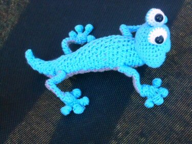 Blauer gehäkelter Amigurumi-Gecko mit großen Augen und vier Beinen auf dunklem Untergrund