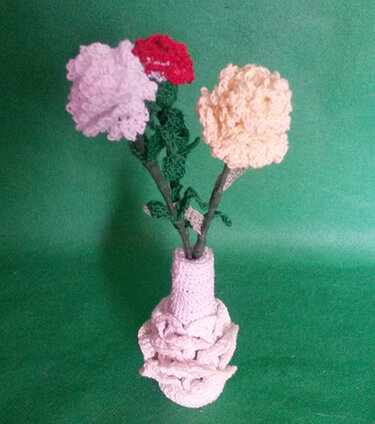 Drei gehäkelte Blumen (rosa, rot, gelb) in einer gehäkelten Vase auf grünem Hintergrund