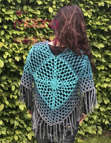 Rückenansicht: gehäkelter Poncho in Türkis‑Grau mit zentralem Blumenmotiv und Fransen, getragen über dunklem Oberteil