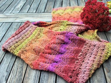 Gestrickter Schal oder Loop mit Farbverlauf in Herbsttönen und unterschiedlichen Strukturmustern auf Holztisch, neben getrockneten roten Blüten.