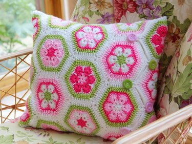 Handgehäkelter Kissenbezug mit sechseckigen African‑Flower‑Motiven in Rosa, Grün und Weiß auf einem Korbstuhl