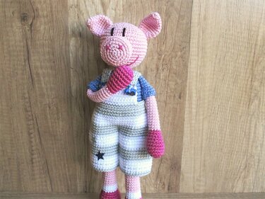 Gehäkeltes rosa Amigurumi-Schwein mit gestreifter Latzhose vor Holz