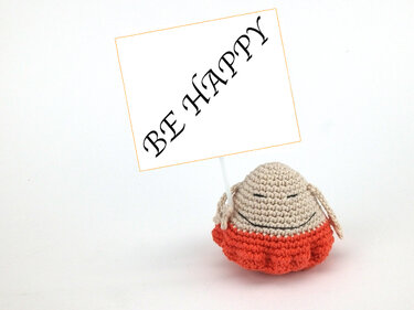 Beiges gehäkeltes Amigurumi mit orangefarbenem Rock und Schild 'BE HAPPY' auf weißem Hintergrund