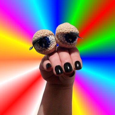 Hand mit schwarzen Nägeln präsentiert zwei gehäkelte Amigurumi-Augen-Fingerpuppen mit blauer Iris und Wimpern