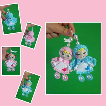 Zwei gehäkelte Amigurumi-Engelchen, eines rosa mit Herz, eines hellblau mit Schmetterling, beide mit Perlen als Beine und Aufhängung, an einer Hand gehalten vor grünem Hintergrund.